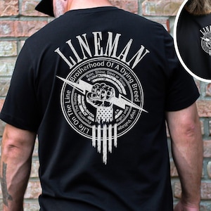 May include: Black t-shirt with a white graphic design on the back. The graphic features a fist holding a lightning bolt with the text "Lineman Brotherhood of a Dying Breed" and "Life on the Line".