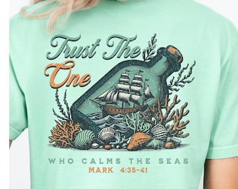 Könnte beinhalten: Mintgrünes T-Shirt mit Grafik auf Vorder- und Rückseite. Die Grafik auf der Vorderseite zeigt "Trust The One" mit einem Schiff in einer Flasche. Die Grafik auf der Rückseite wiederholt den Text und das Design mit dem Text "Who Calms The Seas" und "Mark 4:38-41".