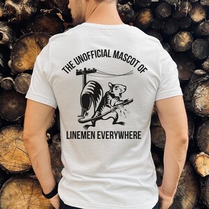 May include: White t-shirt with a black graphic on the back. The graphic features a cartoon rat playing a guitar near a utility pole, with the text "THE UNOFFICIAL MASCOT OF LINEMEN EVERYWHERE".