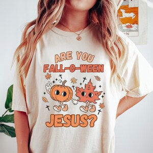 May include: A white t-shirt with the text "Are you Fall-O-Ween Jesus?" in orange and brown lettering. The text is accompanied by a cartoon pumpkin and leaf holding hands.