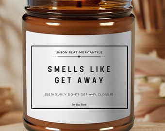 May include: A brown glass candle with a white label that reads "UNION FLAT MERCANTILE", "SMELLS LIKE GET AWAY", "(SERIOUSLY DON'T GET ANY CLOSER)", and "Say Wax Blend".