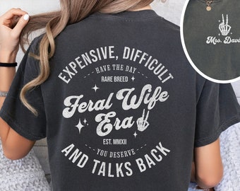 May include: Dark gray t-shirt with white text: "EXPENSIVE, DIFFICULT, FERAL WIFE ERA, AND TALKS BACK." The shirt also has a small image of a skeleton hand making a peace sign. The text "Mrs. Davis" is printed below the skeleton hand.
