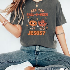 May include: A grey t-shirt with a graphic of a pumpkin and a leaf holding hands. The text on the shirt reads "Are you Fall-O-Ween Jesus?"