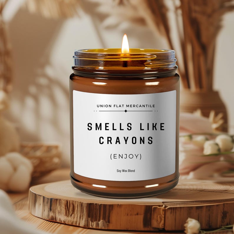 May include: A brown glass candle with a white label that reads "Smells Like Crayons (Enjoy)" Soy Wax Blend. The candle is lit and the flame is visible.