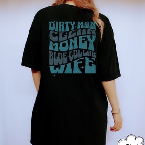 Może przedstawiać: Czarny T-shirt z niebieskim nadrukiem, na kt&oacute;rym widnieje napis "Dirty Man Clean Money Blue Collar Wife" w retro stylu.