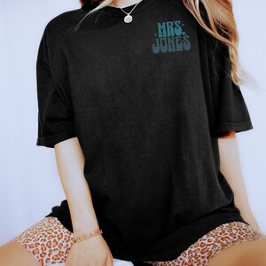 Może przedstawiać: Czarny T-shirt z napisem "MRS. JONES" w retro czcionce.