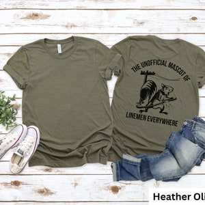 May include: Heather olive-colored t-shirt with a crew neck. The back of the shirt features a black graphic of a squirrel with the text "THE UNOFFICIAL MASCOT OF LINEMEN EVERYWHERE". A pair of blue jeans and white sneakers are also in the image.