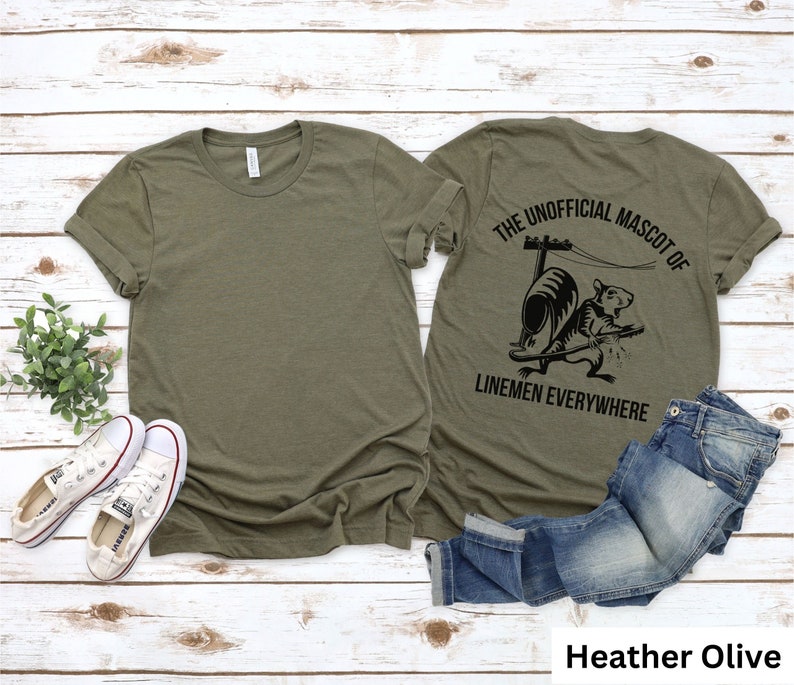 May include: Heather olive-colored t-shirt with a crew neck. The back of the shirt features a black graphic of a squirrel with the text "THE UNOFFICIAL MASCOT OF LINEMEN EVERYWHERE". A pair of blue jeans and white sneakers are also in the image.