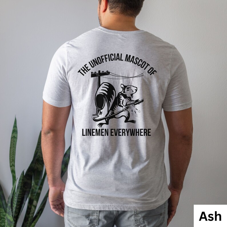 May include: Ash-colored t-shirt with a black graphic on the back. The graphic features a squirrel holding a tool with the text "THE UNOFFICIAL MASCOT OF LINEMEN EVERYWHERE". The shirt is a crew neck and short-sleeved.