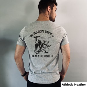 May include: Athletic heather gray t-shirt with a black graphic of a rat holding a tool, a utility pole, and the text "THE UNOFFICIAL MASCOT OF LINEMEN EVERYWHERE". The shirt is short-sleeved.