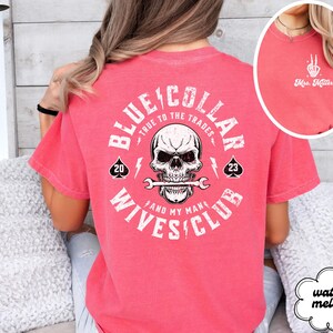 May include: A pink t-shirt with a white graphic on the back that says "Blue Collar True to the Trades and My Man's Wives Club". The graphic includes a skull with a wrench in its mouth and two playing card symbols.