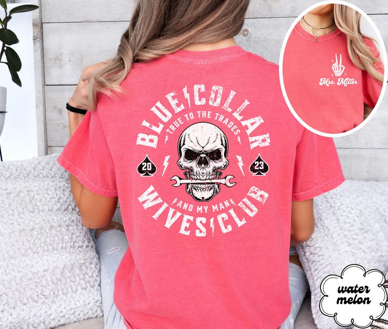 May include: A pink t-shirt with a white graphic on the back that says "Blue Collar True to the Trades and My Man's Wives Club". The graphic includes a skull with a wrench in its mouth and two playing card symbols.