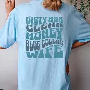Może przedstawiać: Jasnoniebieska koszulka z grafiką w stylu retro, na kt&oacute;rej widnieje napis "Dirty Man Clean Money Blue Collar Wife" w kolorze turkusowym.
