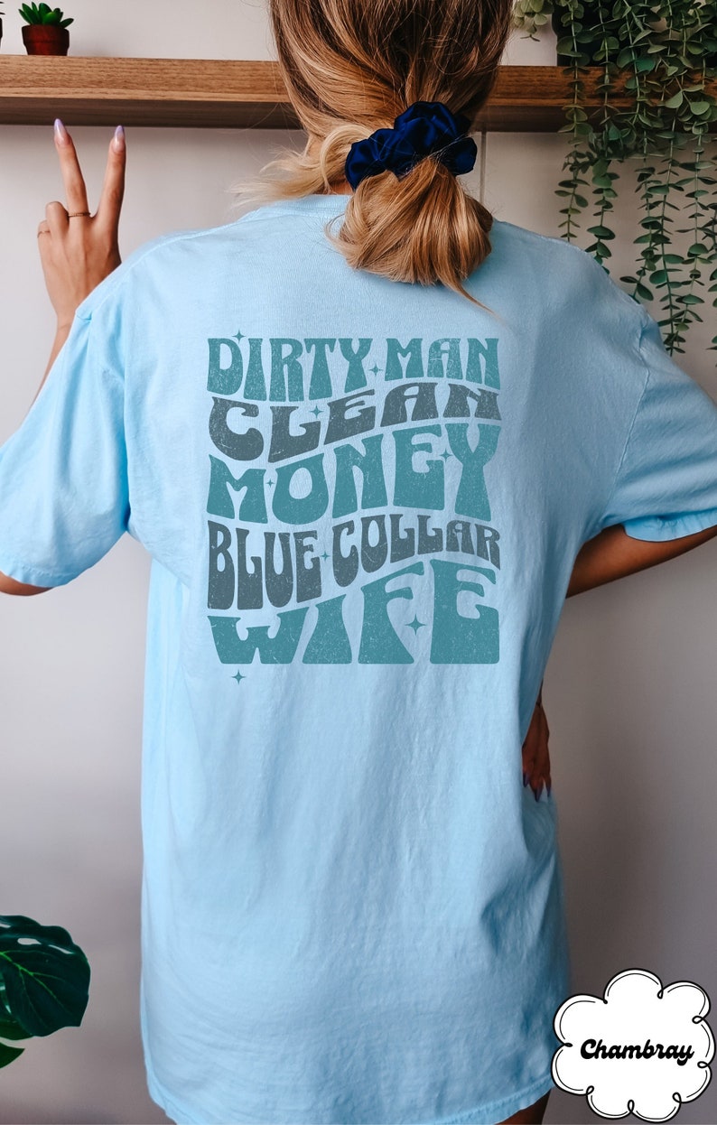 Może przedstawiać: Jasnoniebieska koszulka z grafiką w stylu retro, na kt&oacute;rej widnieje napis "Dirty Man Clean Money Blue Collar Wife" w kolorze turkusowym.