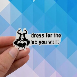 May include: A white sticker with a black illustration of a woman with horns and the text "dress for the job you want". The woman is wearing a black corset and choker. The sticker is held by a hand.