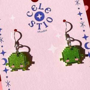 May include: A pair of pixelated green alien earrings with silver hooks are displayed on a pink card. The card has a border of stars, moons, and dots, with the text "Celestio Studio". The earrings have a glittery finish.