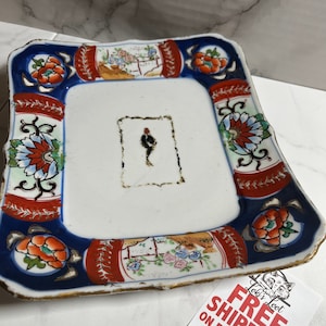 May include: Square ceramic plate with a white background and a gold rim. The plate is decorated with red, blue, and gold floral designs. There is a small, rectangular, white area in the center of the plate with a faded black design.