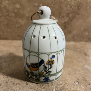 May include: A white ceramic birdcage ornament with a green and gold design. The cage has a small bird with blue, yellow, and orange feathers inside. The ornament has a gold string for hanging.
