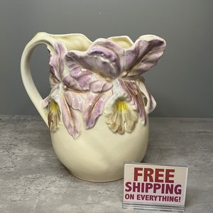 May include: A white ceramic pitcher with a floral design. The pitcher features three large orchid flowers in shades of purple and yellow. The pitcher has a wide mouth and a handle.