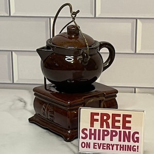 May include: A brown ceramic teapot with a lid and a metal handle. The teapot is sitting on a brown ceramic base with a decorative design. The teapot is on a white tiled surface with a sign that says "FREE SHIPPING ON EVERYTHING!"