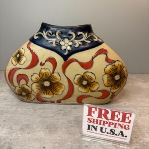 May include: Cream ceramic vase with a floral and ribbon design. The vase has yellow flowers with brown centres and red ribbons against a cream background. The top is navy blue with a floral relief.