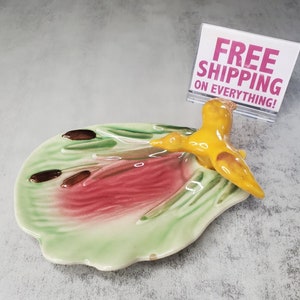 Puede incluir: Un plato de cerámica verde y rosa con una figurita de pájaro amarillo en la parte superior. El plato tiene forma de hoja y tiene un diseño en relieve de juncos y nenúfares. El pájaro está posado en el borde del plato y mira hacia la derecha.