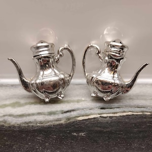 May include: A pair of silver teapot-shaped salt and pepper shakers. The shakers have a detailed design and are made of metal.