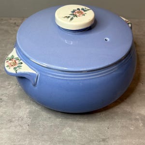 May include: A light blue ceramic casserole dish with a lid. The lid features a white knob with a floral design in pink, green, and blue. The handles also have floral accents. A small hole is visible on the lid.