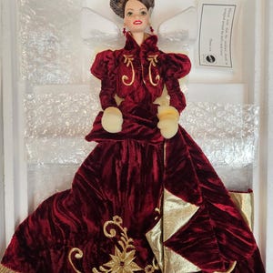 Puede incluir: Muñeca con un vestido de terciopelo burdeos con bordados y adornos dorados. La muñeca tiene una chaqueta a juego con detalles dorados y guantes blancos. El pelo de la muñeca está peinado en un moño y lleva pendientes colgantes. La muñeca está en una caja blanca.