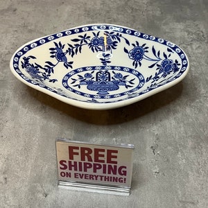 May include: A white ceramic bowl with a blue and white floral design. The bowl is square shaped with rounded edges. The bowl is sitting on a grey surface. The text "FREE SHIPPING ON EVERYTHING!" is on a sign in front of the bowl.
