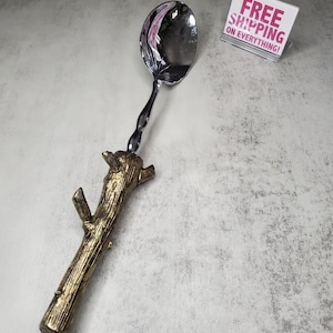 May include: A silver spoon with a gold-painted tree branch handle. The spoon is a serving spoon with a rounded bowl. The handle is a realistic-looking tree branch with a textured surface.