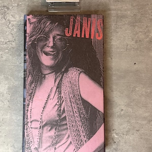 Puede incluir: Un libro con una portada rosa y marrón que presenta una imagen en blanco y negro de Janis Joplin. La palabra "JANIS" está en letras rojas grandes. La imagen muestra a una mujer sonriendo con gafas de sol.