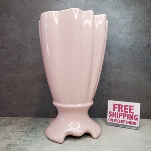 Könnte beinhalten: Eine hellrosa Keramikvase mit geriffeltem Design und einem ausgestellten Fuß. Die Vase hat eine einzigartige, wellenförmige Form und eine glänzende Oberfläche.
