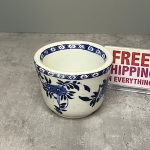 May include: A small white ceramic cup with a blue and white floral pattern. The cup has a scalloped rim and a blue and white geometric pattern around the top.