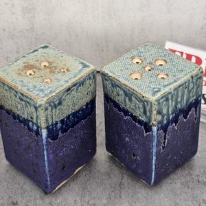 May include: Two square salt and pepper shakers featuring a dual-tone glaze. The top sections are a light, textured sage green with five small holes, while the bottom sections are a deep, vibrant purple with a rough, speckled finish.