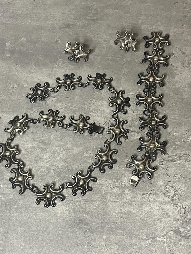 Peut inclure: Un ensemble de collier, bracelet et boucles d'oreilles en argent avec un motif g&eacute;om&eacute;trique. Les bijoux sont faits de maillons en argent entrelac&eacute;s avec un cercle central sur&eacute;lev&eacute; sur chaque maillon.