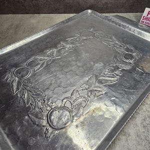 May include: A rectangular, silver-coloured metal serving tray. The tray features a raised, embossed design of fruit and leaves around the centre. The tray has a slightly textured surface and a decorative edge. The tray is likely vintage.