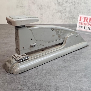 Puede incluir: Una grapadora gris vintage con una base curva y una parte superior rectangular. La grapadora muestra signos de desgaste, con arañazos visibles y pérdida de pintura. La grapadora de metal está diseñada para uso en la oficina.