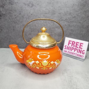 May include: Orange ceramic teapot with a gold lid and handle. The teapot features a floral design with white birds and yellow flowers.