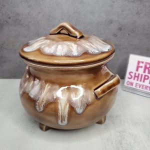 以下が含まれることがあります： 白釉のアクセントが施された茶色の陶器製の鍋。鍋には2つの取っ手と3つの小さな足があります。