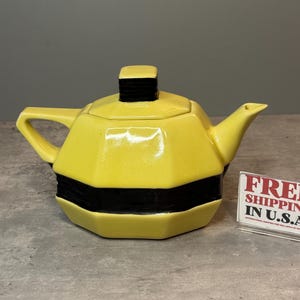 Könnte beinhalten: Eine gelbe Keramik-Teekanne mit einem schwarzen Streifen in der Mitte und einem schwarzen quadratischen Griff. Die Teekanne hat einen Ausguss und einen Griff. Die Teekanne steht auf einer grauen Oberfläche.