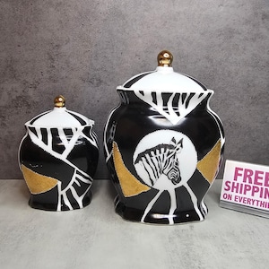 Könnte beinhalten: Zwei dekorative Keramikbehälter mit Deckeln. Die Behälter sind schwarz mit weißen und goldenen Akzenten und zeigen ein Zebra-Kopf-Design. Die Deckel sind weiß mit schwarzen Streifen und goldenen Knäufen. Die Behälter sind unterschiedlich groß.