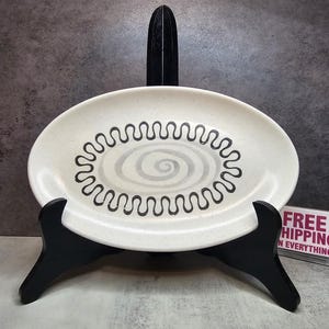 May include: Oval ceramic serving dish with a cream-colored base and a black, wavy spiral design. The dish is displayed on a black wooden stand. The dish has a speckled texture and a minimalist aesthetic.