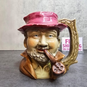 Könnte beinhalten: Ein Keramikbecher in Form eines Männerkopfes, der einen rosa Hut trägt und eine Pfeife raucht. Der Becher hat einen Henkel und ist mit realistischen Details bemalt, darunter ein Bart und ein brauner Mantel.