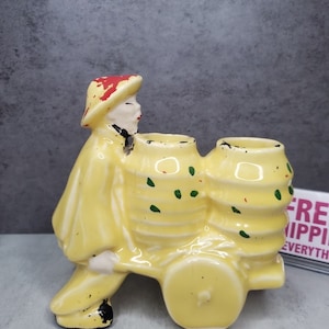 Puede incluir: Un macetero de cerámica amarillo con forma de persona empujando un carro de mano con dos barriles. La persona lleva un sombrero rojo y amarillo y una chaqueta amarilla. Los barriles tienen detalles pintados en verde y rojo.