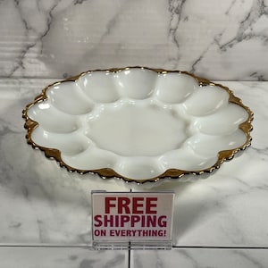 Puede incluir: Plato para huevos de vidrio blanco lechoso con borde dorado. El plato tiene 6 secciones individuales para huevos. El plato está sobre una superficie de mármol con un cartel que dice "¡ENVÍO GRATIS EN TODO!"