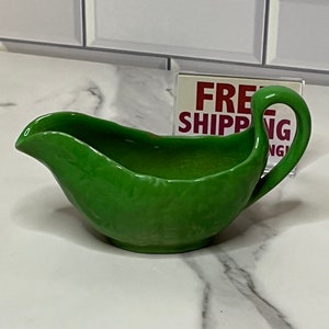 May include: A green ceramic gravy boat with a textured surface. The boat has a handle on the right side and a spout on the left side.