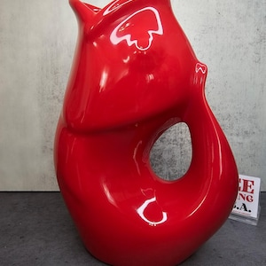 May include: A glossy, red ceramic pitcher with a unique, organic shape. The pitcher features a large, open handle and a spout. The surface reflects light, highlighting its curves and smooth finish. The design is modern and sculptural.