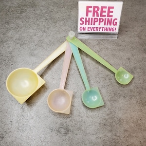 Puede incluir: Conjunto de cuatro cucharas medidoras de color pastel. Las cucharas son de plástico y tienen una forma redondeada. Las cucharas están dispuestas en forma de abanico sobre una superficie gris. Las cucharas son amarillas, rosas, verdes y azules.