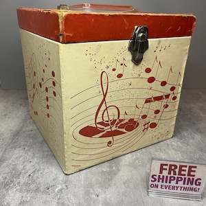 May include: A vintage record storage box with a red lid and a white base. The box is decorated with a musical note design and a red record with a treble clef. The box has a metal latch.